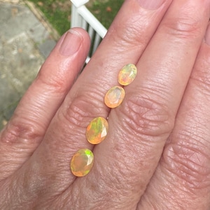 May include: Four oval-shaped, faceted opals with a translucent, warm orange base color and iridescent flashes of green and yellow light. The gemstones are arranged in a vertical line, each displaying a polished surface and a multi-faceted cut.
