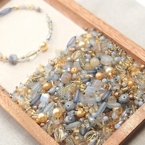 Peut inclure: Un plateau en bois rempli d'un assortiment de perles dans les tons or, gris et transparent. Un bracelet de perles avec des perles dorées et grises est dans le coin supérieur gauche. Les perles varient en forme et en taille.