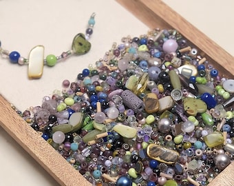 May include: A wooden tray filled with a variety of colorful beads in shades of blue, green, purple, and pearl. A partially assembled necklace with a heart-shaped bead and a white rectangular bead is visible. The beads are of various shapes and sizes.