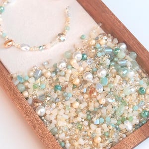 Op de afbeelding: Een houten bakje gevuld met een assortiment kralen in verschillende vormen, maten en kleuren, waaronder wit, crème, goud en tinten blauw en groen. Een kralenarmband met vergelijkbare kleuren rust op een wit oppervlak.