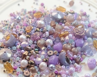 May include: A collection of assorted beads in shades of purple, lavender, gold, and pearl. The beads vary in size and shape, including round, faceted, and decorative elements like moons and roses. Ideal for jewelry making and crafting.