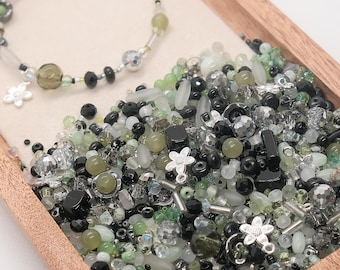 May include: A wooden tray filled with an assortment of beads in shades of green, black, and clear. A completed bracelet with flower charms sits on top. The beads vary in shape and size, including round, faceted, and oblong.