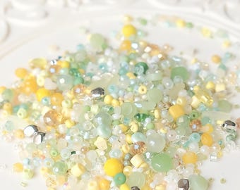 May include: A collection of assorted beads in shades of yellow, green, and clear. The beads vary in size and shape, including round, square, and faceted designs. Some beads have a metallic sheen, and the arrangement is scattered on a white surface.