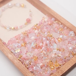 Peut inclure: Un plateau en bois rempli d'un assortiment de perles roses, blanches et dorées, de cristaux et de petits éléments décoratifs. Un bracelet délicat avec des perles roses et des accents dorés repose sur une surface claire à l'intérieur du plateau. Les perles varient en taille et en forme.
