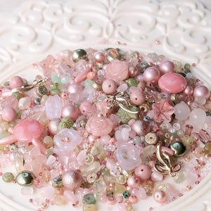 May include: A collection of assorted craft beads in shades of pink, rose, and pale green. The mix includes round pearls, faceted crystals, translucent flower-shaped beads, rose-shaped beads, and small metallic charms in a silver-toned finish.