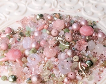 May include: A collection of assorted craft beads in shades of pink, rose, and pale green. The mix includes round pearls, faceted crystals, translucent flower-shaped beads, rose-shaped beads, and small metallic charms in a silver-toned finish.