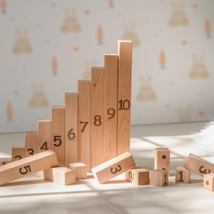 May include: Wooden number blocks arranged in a staircase, with numbers 1 through 10. Some blocks are scattered around the base. The blocks are made of light-colored wood. The background features a bunny and carrot pattern.