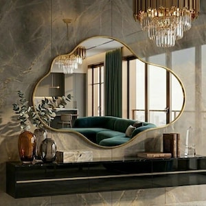 May include: An irregular, cloud-shaped wall mirror with a thin gold frame, mounted above a long, black, wall-mounted floating console cabinet with horizontal gold trim, set against a large-format, polished, grey marble-patterned wall panel.