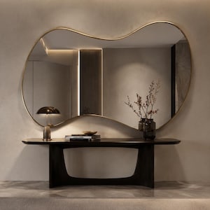 May include: A large, irregular, organic-shaped wall mirror with a thin gold frame mounted above a dark brown wooden console table. The table features a curved base and holds a small lamp, a stack of books, and a glass vase with thin branches.