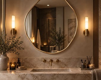 May include: A round mirror with a thin gold frame mounted on a wall above a marble vanity. The vanity features a gold faucet, two gold wall sconces with glowing lights, a gray vase with greenery, and various bathroom accessories.