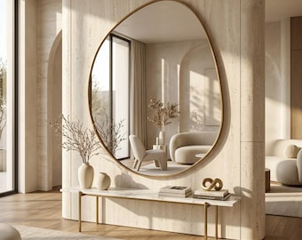May include: A large, organic-shaped wall mirror with a thin gold frame mounted on a beige stone wall above a white marble console table with gold legs. The table holds two ceramic vases with branches and a stack of books with a gold sculpture.
