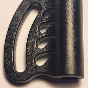 May include: Black plastic buckle with a single hole and a series of four slots. The buckle is designed to be used with a strap or cord.