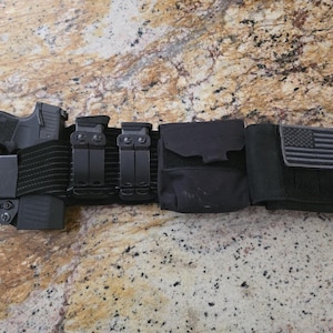 May include: A black tactical belt with a handgun holster, two magazine pouches, a utility pouch, and a pouch with a small American flag patch.