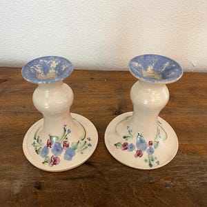 May include: A pair of white ceramic candle holders with blue and pink floral designs. The holders have a flared top and a round base.