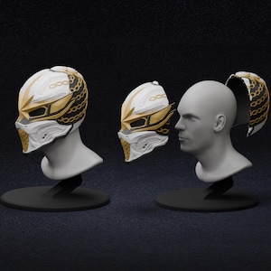 K&ouml;nnte beinhalten: Ein wei&szlig;-goldener Helm mit einem schwarzen Seitenteil, das ein goldenes Kettenmuster aufweist. Der Helm hat ein sternf&ouml;rmiges Detail auf der Vorderseite und einen perforierten goldenen Mundschutz. Er wird auf einer B&uuml;ste montiert und zerlegt gezeigt.