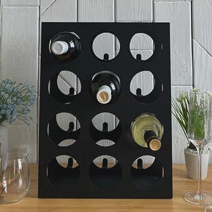 Może przedstawiać: Czarny stojak na wino z okrągłymi otworami mieści kilka butelek wina. Stojak stoi na drewnianej powierzchni, w pobliżu kieliszki do wina i butelki ginger ale. Półka nad nim prezentuje elementy dekoracyjne i roślinę doniczkową na białej ścianie.