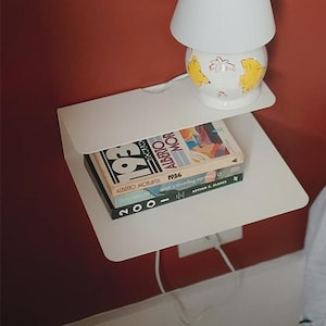 Könnte beinhalten: Eine weiße Nachttischlampe mit weißem Lampenschirm und floralem Design auf dem Sockel steht auf einem weißen Regal. Unter der Lampe befinden sich zwei Bücher. Das Regal ist an einer roten Wand neben einem Bett mit weißer Bettwäsche befestigt.