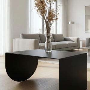 Op de afbeelding: Een matzwarte salontafel met een plat rechthoekig blad en twee gebogen, halfronde zijsteunen. In het midden van de tafel staat een heldere glazen vaas met een hoog arrangement van gedroogde beige grassen en tarwestengels.