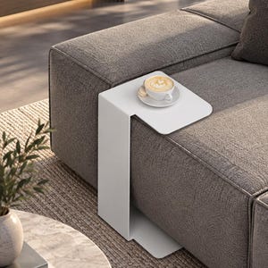 May include: A white C-shaped side table with a smooth, matte finish, designed to slide over the edge of a sofa. The table features a flat, rectangular top surface and a vertical support base that extends to the floor, holding a white coffee cup.