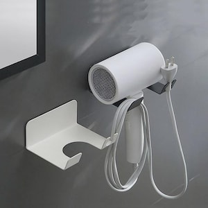 May include: A white hair dryer and its holder are mounted on a gray wall. The hair dryer is white with a black mesh front and a gray cord. The holder is white and designed to hold the hair dryer and its cord.
