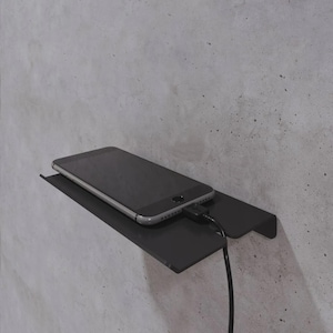 May include: A black smartphone rests on a black wall-mounted shelf, connected to a charging cable. The shelf is made of metal and is attached to a gray concrete wall. The phone is silver and black.