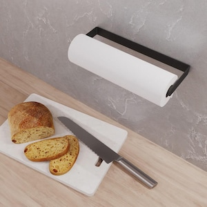 May include: A matte black metal paper towel holder mounted horizontally, holding a roll of white paper towels. Below, a white rectangular cutting board holds a loaf of crusty bread and two slices, alongside a serrated bread knife with a silver blade.