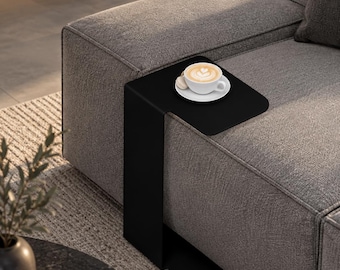 May include: A matte black C-shaped side table with a flat top surface and a vertical support base that slides under a sofa. The table features a smooth, solid metal construction and is positioned to hold a white ceramic cup and saucer.