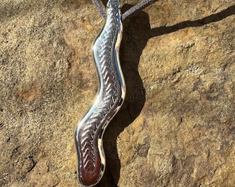 Op de afbeelding: Een zilveren ketting met een golvende, getextureerde hanger. De hanger is bevestigd aan een bruin leren koord.