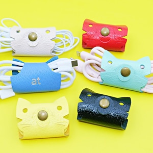 May include: A collection of colorful, cat-shaped cable organizers. The organizers are in various colors, including pink, red, blue, yellow, and black. Each organizer has a snap closure and holds white cables. The blue organizer has the letters "at" on it.