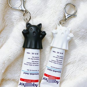 May include: Two lip balm tubes with keychain attachments, each featuring a ghost-shaped cap with cat ears. One cap is solid black and the other is solid white. The tubes are labeled with text including 'Immediate Relief', 'LIP REPAIR', and '35 FL OZ 10ML'.