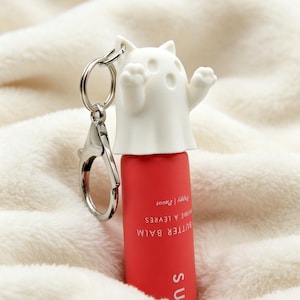 May include: A red cylindrical lip balm tube with a white silicone ghost-shaped cap featuring cat ears and raised arms. A silver metal keychain ring is attached to the top of the ghost cap. The tube is printed with the text BUTTER BALM, BAUME À LÈVRES, and Poppy | Pavot.