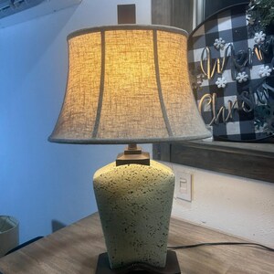 May include: A green ceramic table lamp with a beige fabric lampshade. The lamp has a textured finish and a brown base.