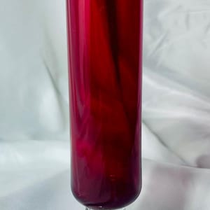 May include: A tall, ruby red glass with a clear, decorative stem and base. The glass has a long, cylindrical shape and is likely a champagne flute or similar stemware. The glass is approximately 10 inches tall.