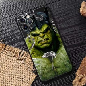 Op de afbeelding: Zwarte telefoonhoes met de Hulk van Marvel Comics. De groenhuidige superheld wordt afgebeeld met een boze uitdrukking, die door een gebarsten glaseffect breekt. De hoes heeft een glanzende afwerking en een camera-uitsparing.