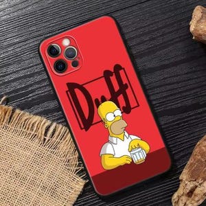 Può includere: Custodia per telefono rossa con bordo nero, raffigurante Homer Simpson con una birra e la scritta "Duff" in nero. La custodia ha un modulo fotocamera.