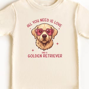 Puede incluir: Una camiseta de color natural con un gr&aacute;fico de un Golden Retriever con gafas de sol en forma de coraz&oacute;n. El texto dice "ALL YOU NEED IS LOVE AND A GOLDEN RETRIEVER." La camiseta tiene mangas cortas y cuello redondo.