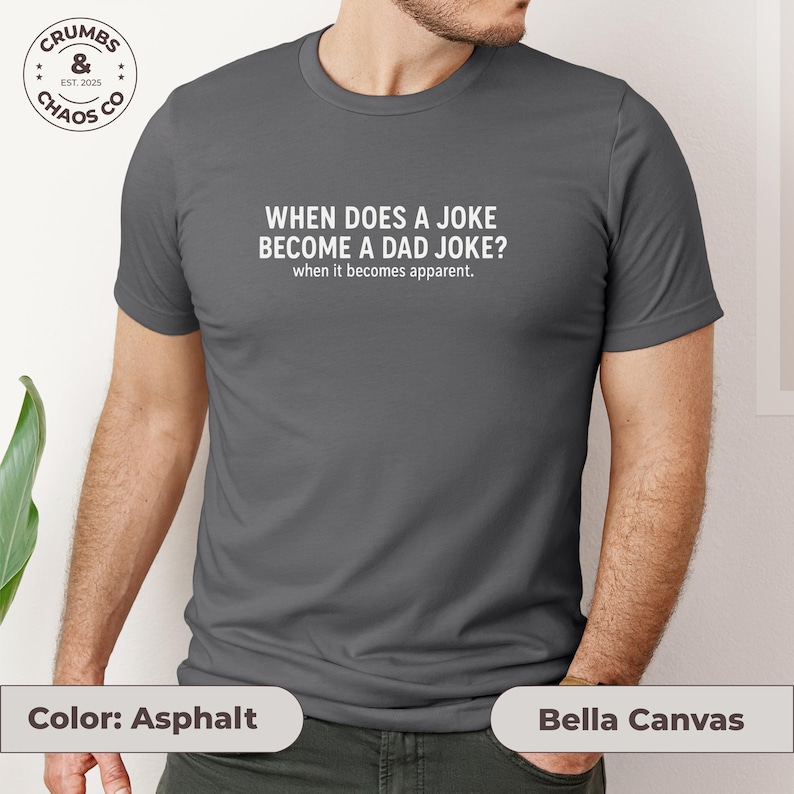 May include: Asphalt-colored Bella Canvas t-shirt with the text "WHEN DOES A JOKE BECOME A DAD JOKE? when it becomes apparent." A circular logo with the text "CRUMBS & CHAOS CO EST. 2025" is visible on the upper left.