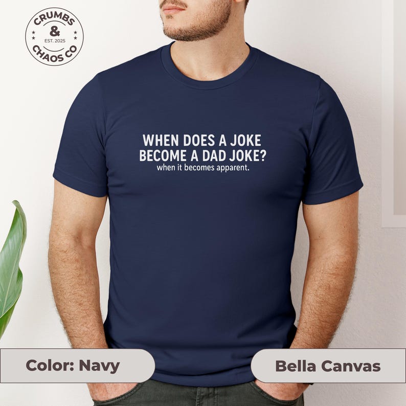 May include: Navy blue t-shirt with the text "WHEN DOES A JOKE BECOME A DAD JOKE? when it becomes apparent." The shirt has a crew neck and short sleeves. The brand logo "CRUMBS & CHAOS CO" is visible in the upper left corner.