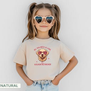 Puede incluir: Una camiseta beige con un gr&aacute;fico de un Golden Retriever con gafas de sol en forma de coraz&oacute;n. El texto "ALL YOU NEED IS LOVE and a GOLDEN RETRIEVER" est&aacute; impreso encima y debajo de la ilustraci&oacute;n del perro. La ni&ntilde;a lleva gafas de sol en forma de coraz&oacute;n.
