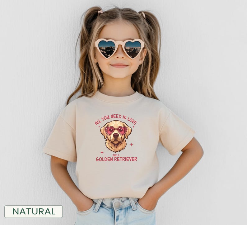 Puede incluir: Una camiseta beige con un gr&aacute;fico de un Golden Retriever con gafas de sol en forma de coraz&oacute;n. El texto "ALL YOU NEED IS LOVE and a GOLDEN RETRIEVER" est&aacute; impreso encima y debajo de la ilustraci&oacute;n del perro. La ni&ntilde;a lleva gafas de sol en forma de coraz&oacute;n.