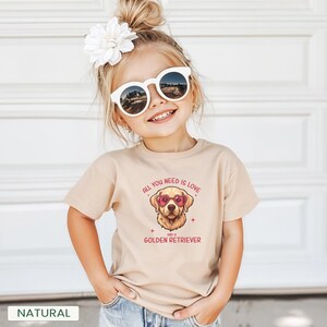 Puede incluir: Una camiseta beige con un gr&aacute;fico de un Golden Retriever con gafas de sol en forma de coraz&oacute;n. El texto en la camiseta dice "ALL YOU NEED IS LOVE AND A GOLDEN RETRIEVER". Un ni&ntilde;o peque&ntilde;o lleva la camiseta, gafas de sol y una flor en el pelo.
