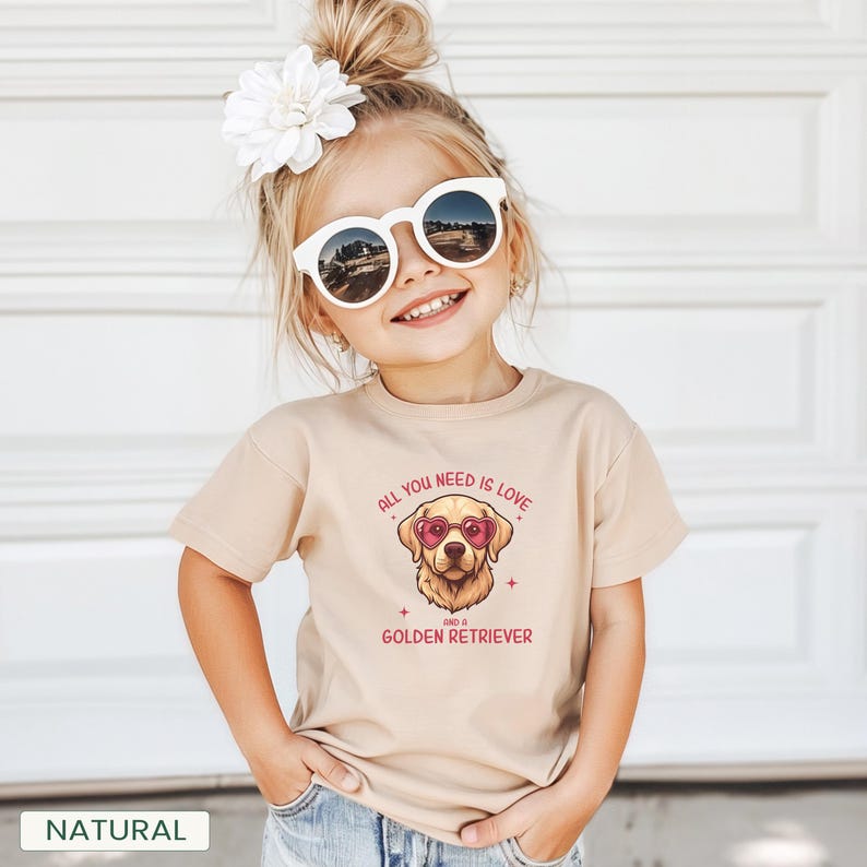 Puede incluir: Una camiseta beige con un gr&aacute;fico de un Golden Retriever con gafas de sol en forma de coraz&oacute;n. El texto en la camiseta dice "ALL YOU NEED IS LOVE AND A GOLDEN RETRIEVER". Un ni&ntilde;o peque&ntilde;o lleva la camiseta, gafas de sol y una flor en el pelo.