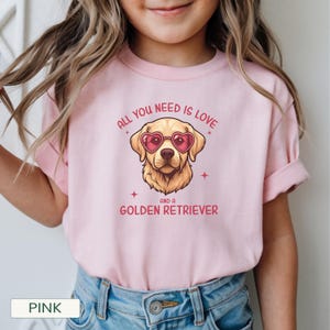 Puede incluir: Camiseta rosa claro con un dise&ntilde;o de Golden Retriever con gafas de sol en forma de coraz&oacute;n. El texto dice "ALL YOU NEED IS LOVE AND A GOLDEN RETRIEVER". La camiseta es de material suave y es adecuada para ni&ntilde;os.