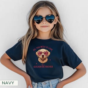 Puede incluir: Camiseta azul marino con un dise&ntilde;o de Golden Retriever con gafas de sol en forma de coraz&oacute;n. El texto "All you need is love and a Golden Retriever" est&aacute; impreso encima y debajo de la ilustraci&oacute;n del perro. La camiseta la lleva una persona.
