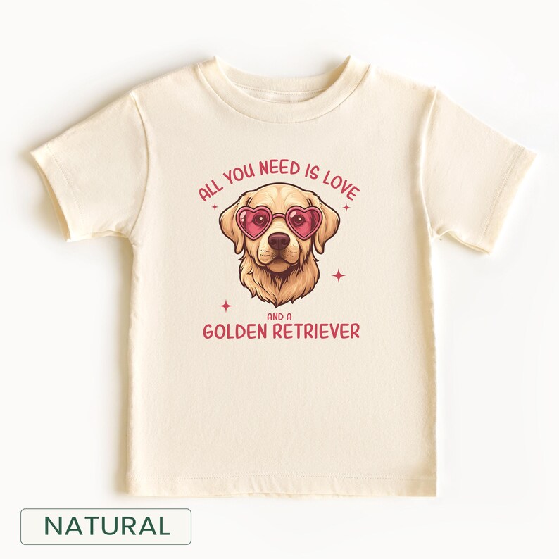 Puede incluir: Una camiseta de color natural con un gr&aacute;fico de un Golden Retriever con gafas de sol en forma de coraz&oacute;n. El texto dice "ALL YOU NEED IS LOVE AND A GOLDEN RETRIEVER." La camiseta tiene mangas cortas y cuello redondo.