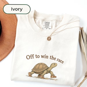 May include: An ivory t-shirt featuring a graphic of a brown tortoise wearing sneakers with the text "Off to win the race" printed in brown above the animal. The shirt has a crew neckline and a Comfort Colors brand label on the inside collar.