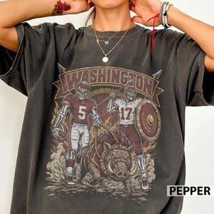 May include: A charcoal gray t-shirt featuring a graphic print of two skeleton football players in burgundy and gold uniforms with the numbers 5 and 17, standing beside a wild boar. The text WASHINGTON is arched above the figures in a banner.