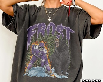May include: Dark gray t-shirt with a graphic featuring the word "FROST" in purple, a hockey player skeleton, and a black bear. The design includes a forest and flames, with the word "PEPPER" in white.