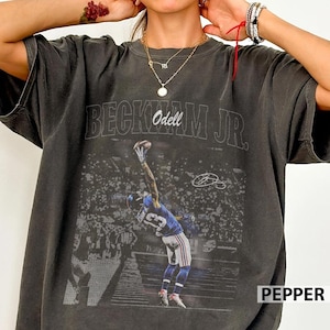 May include: A charcoal gray short-sleeve t-shirt featuring a graphic print of a football player catching a ball. The text "BECKHAM JR." and "Odell" is printed above the image. The shirt has a crew neckline and a relaxed, boxy fit.