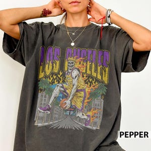May include: Dark gray t-shirt with a graphic of a skeleton basketball player in a yellow and purple uniform, set against a cityscape with the text "LOS ANGELES". The design includes flames and palm trees, creating a vintage, distressed look.